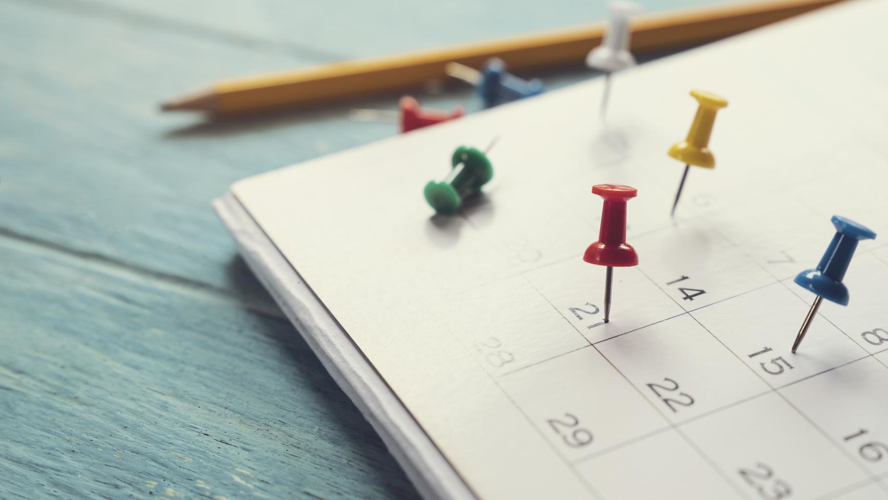 calendar with different color tacks, pencil above calendar, all on table